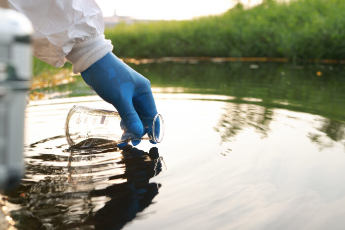 A beaker being dipped into a pond by a gloved hand