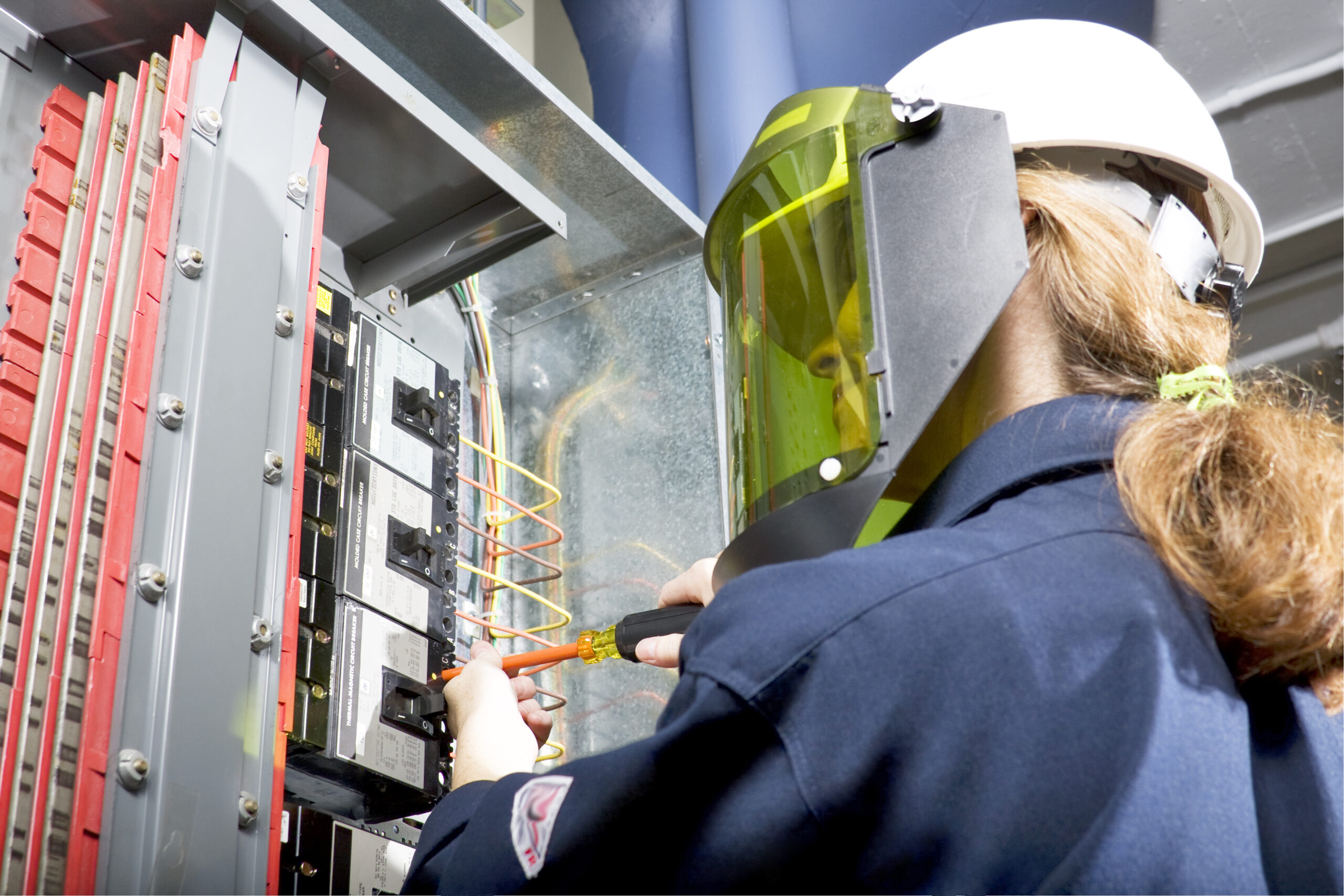 Female electrician with ARC flash protective clothing performing work in an energized panel with a happy smile.