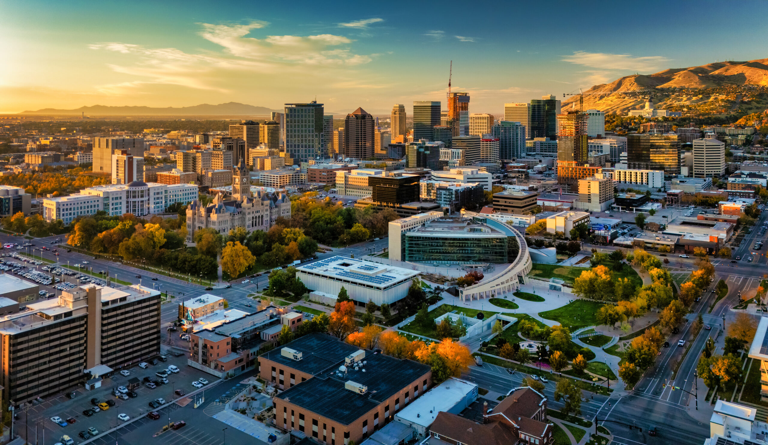 Skyline Aerial of Downtown Salt Lake City during the golden hour / sunset in autumn, including the Utah State Capitol and mountains on the right and various government buildings including the Salt Lake City Public Library (center) in the foreground.